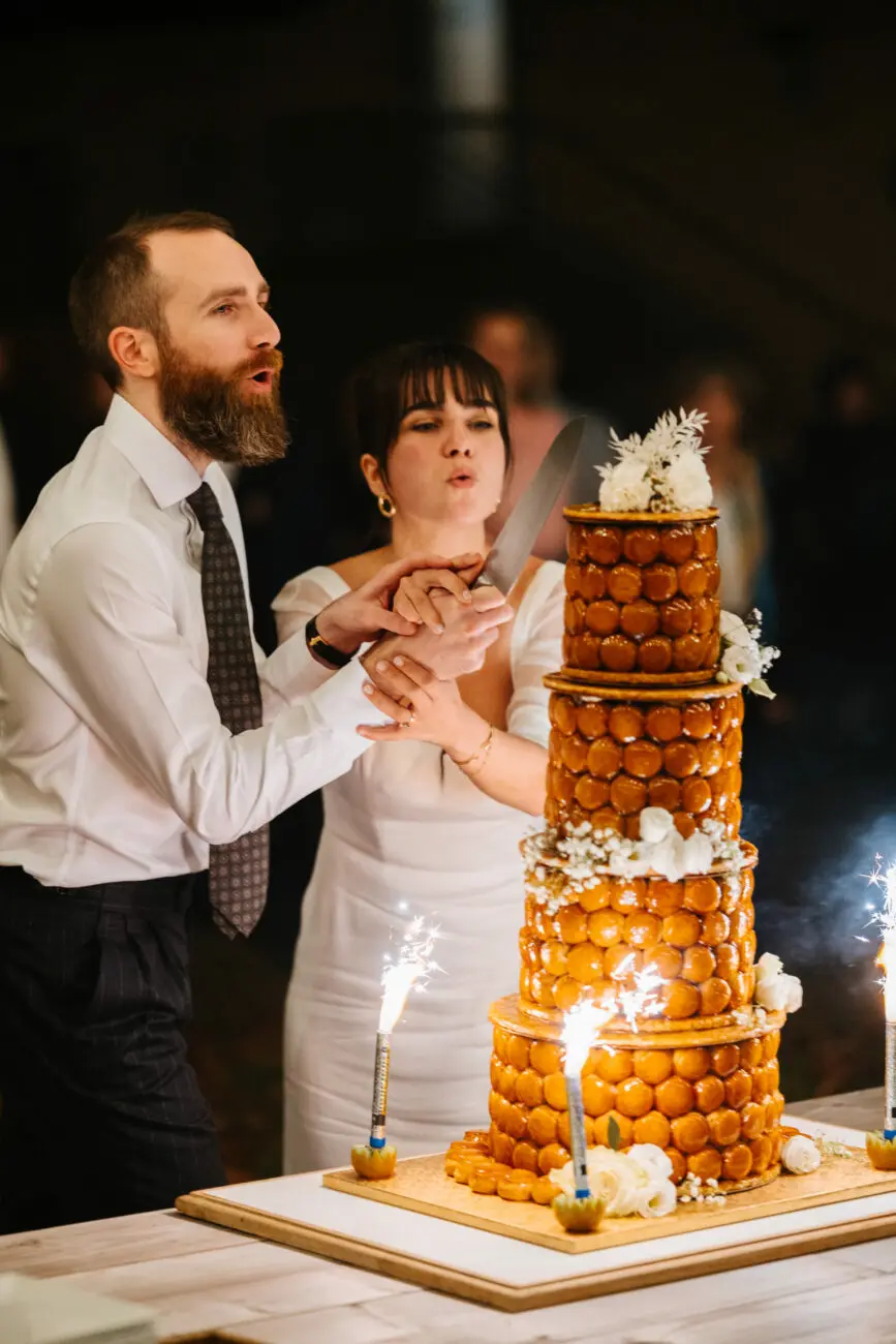 Gros plan d'une mariée et d'un marié découpant ensemble une grande pièce montée de choux traditionnelle, avec des bougies d'anniversaire étincelantes à la base.