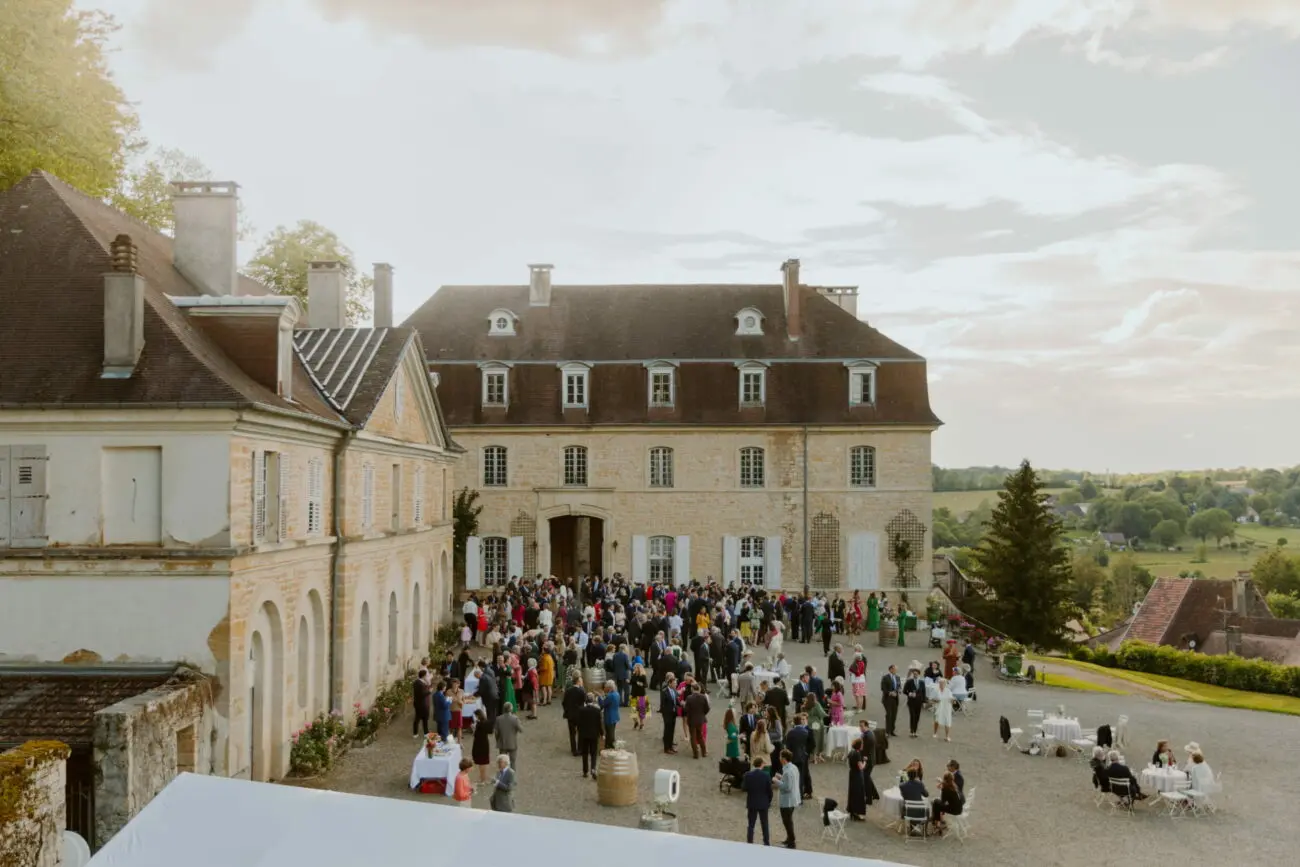 Vue aérienne d'une grande réception de mariage en plein air, avec de nombreux invités en tenue formelle rassemblés dans la cour en gravier d'un château historique en pierre, surplombant un paysage vallonné.