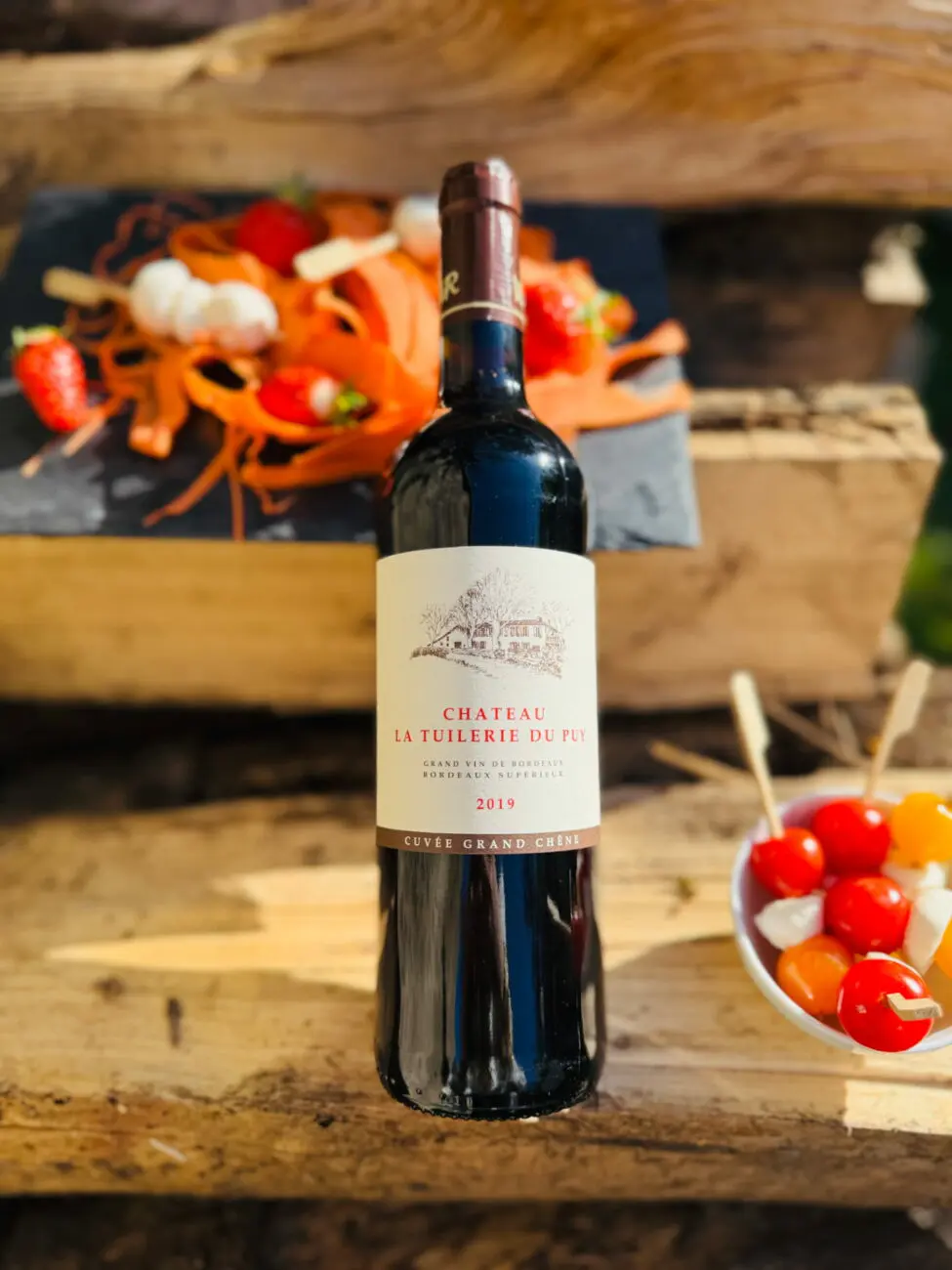 Bouteille de vin rouge Château La Tuilerie Du Puy "Cuvée Grand Chêne" 2019 posée sur des planches en bois lors d'un cocktail de mariage en extérieur, avec des amuses-bouches.
