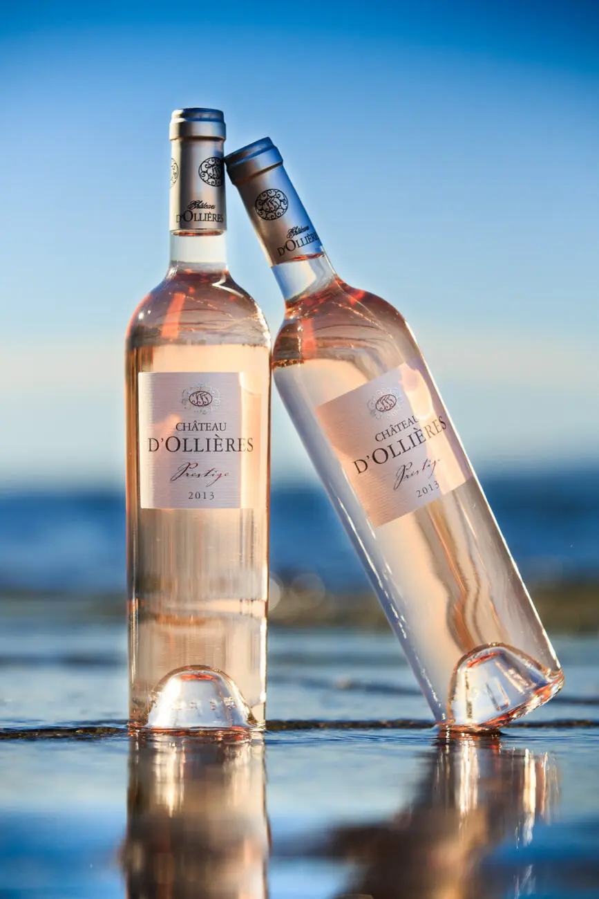 Deux bouteilles de vin rosé Château d'Ollières Prestige posées et se reflétant sur une surface d'eau devant un horizon de mer bleue et ciel bleu.