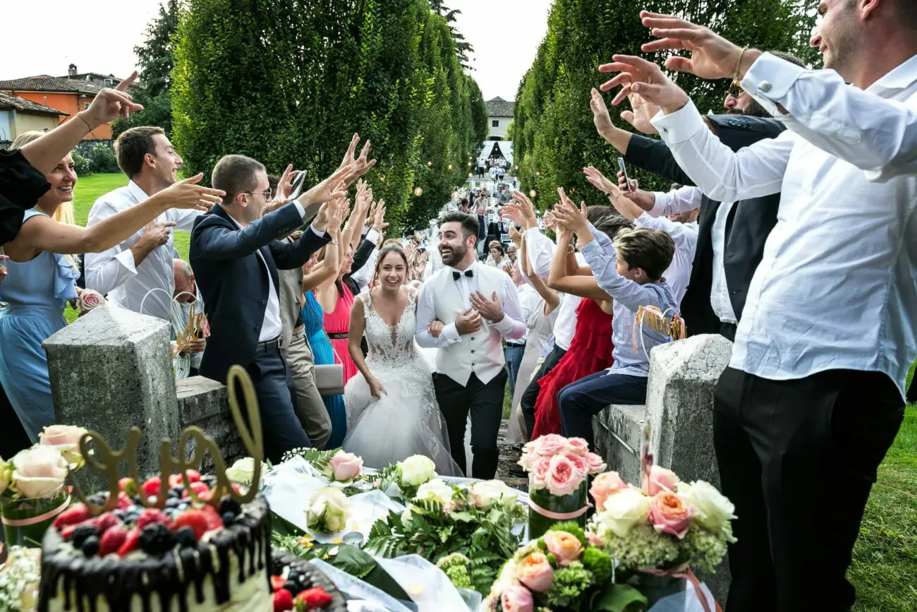 Sortie triomphale de jeunes mariés à travers une haie d'invités joyeux levant les mains, lors d'une réception de mariage en plein air avec buffet de desserts au premier plan.