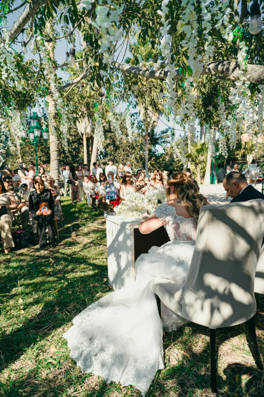 Mariée vue de dos signant un registre lors d'une cérémonie de mariage en plein air sous des arbres décorés de fleurs blanches.
