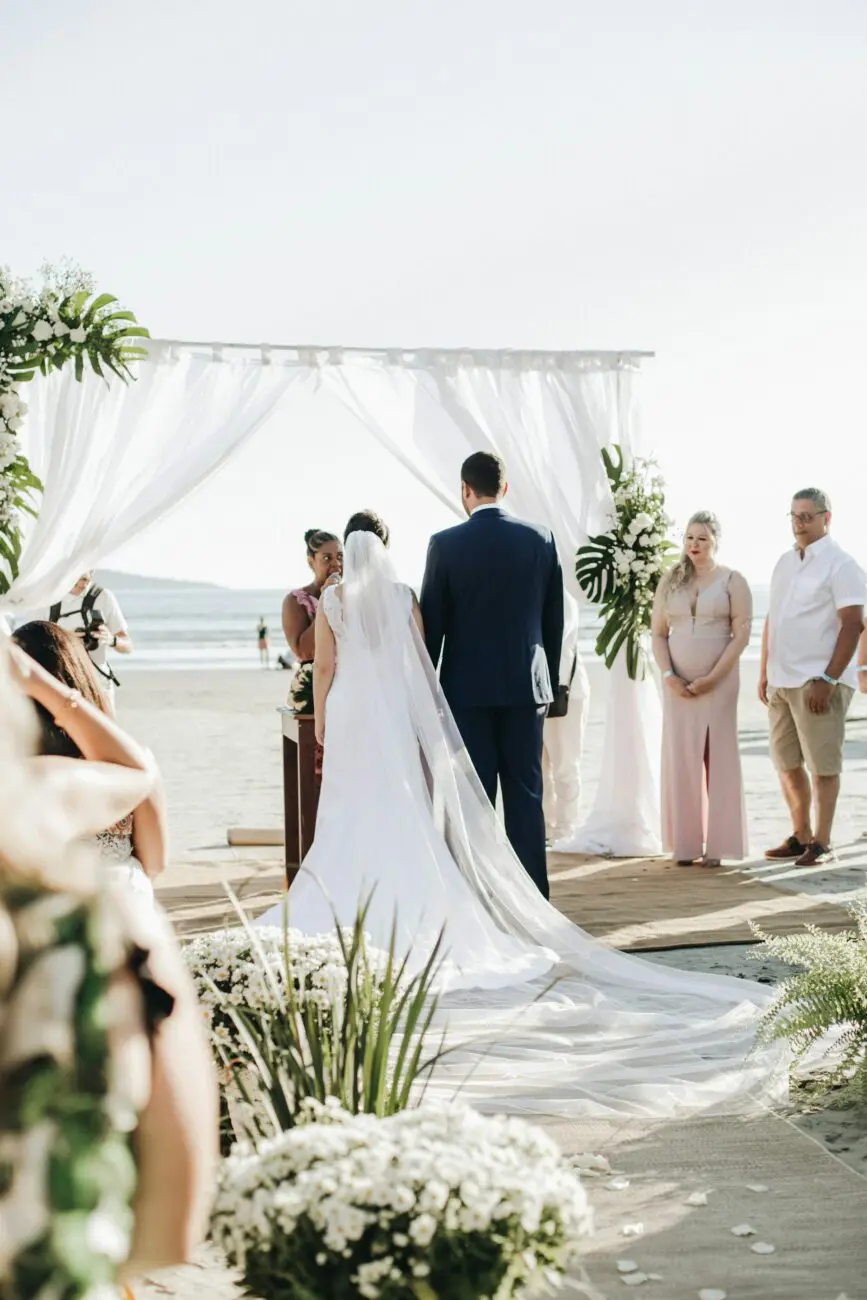 Cérémonie de mariage laïque en extérieur sur la plage avec une arche florale et des invités installés, vue de dos du couple.