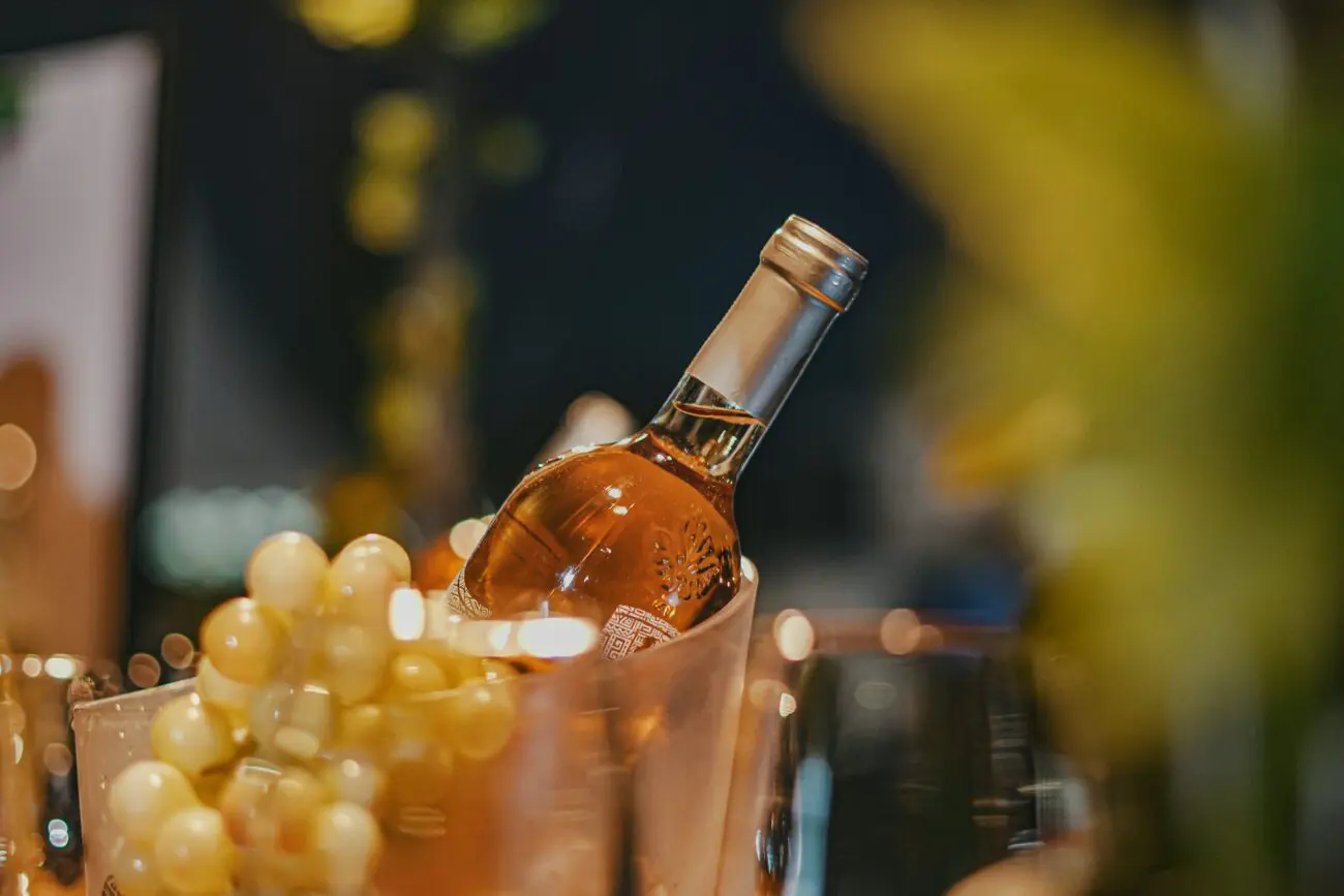 Bouteille de vin rosé dans un seau à glace avec des grains de raisin blanc à côté, dans une ambiance de soirée lumineuse et floue.