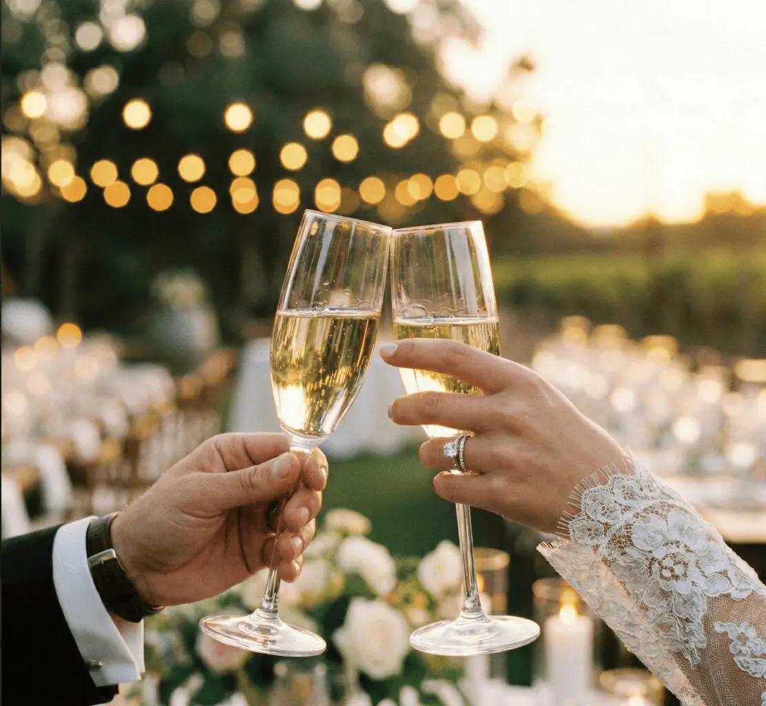 Mains des mariés trinquant avec deux flûtes de champagne lors d'une réception de mariage en extérieur au coucher du soleil.
