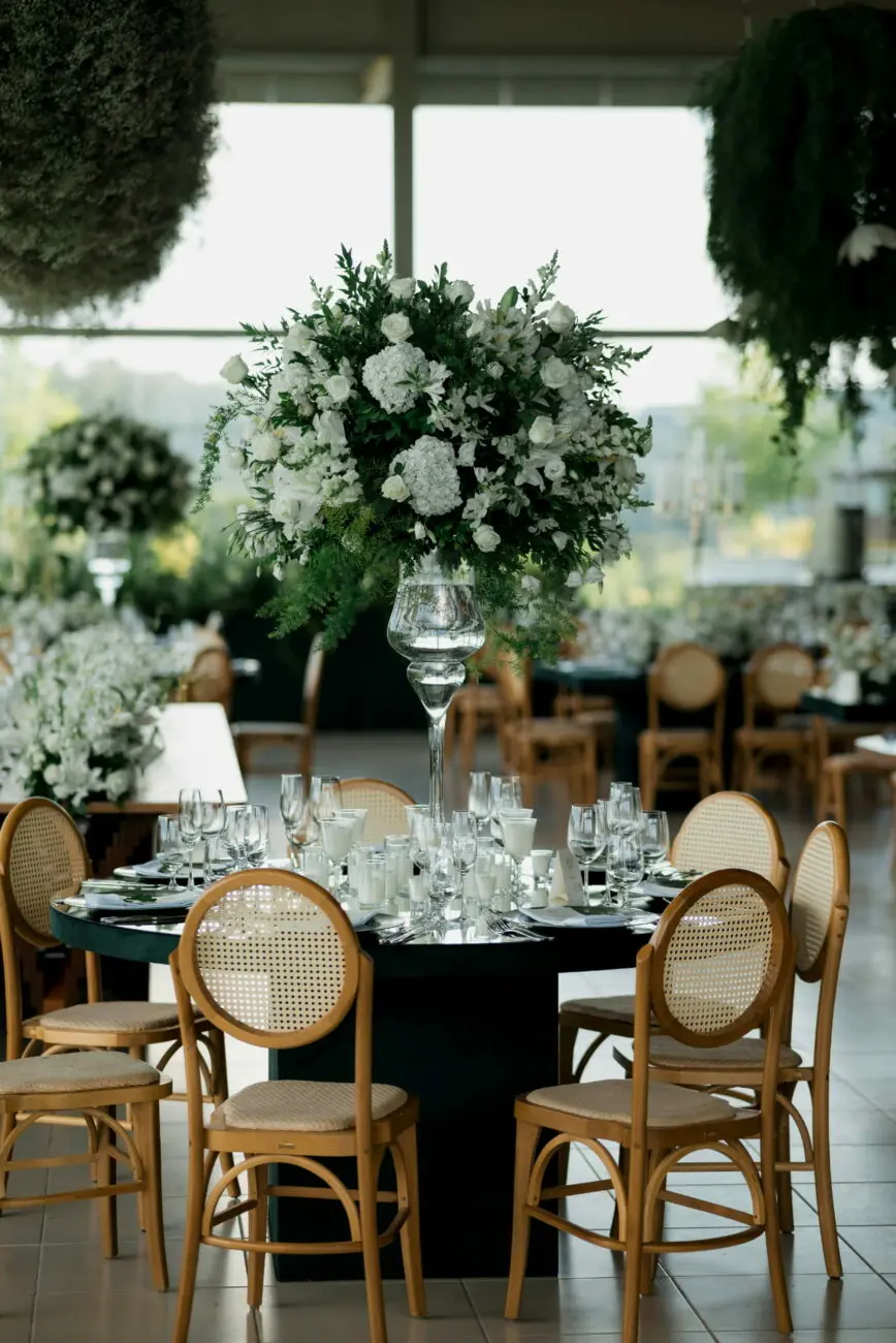 Table de mariage ronde élégante avec chaises en cannage, nappe sombre et grand centre de table floral blanc dans une salle lumineuse.