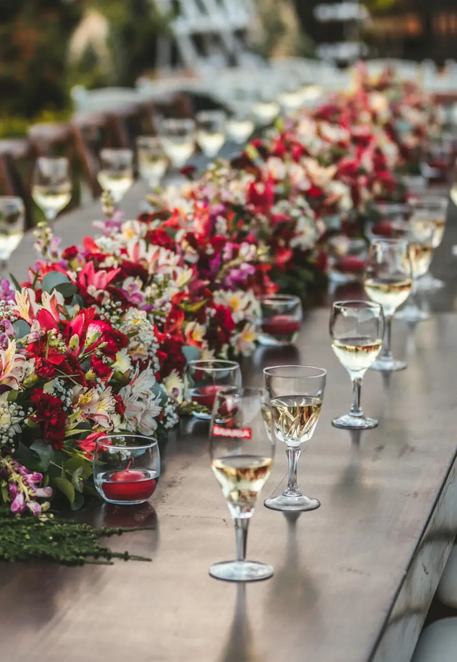 Longue table de mariage en bois sombre ornée d'un chemin de table floral dense aux tons rouges et roses, avec des verres de vin blanc alignés.