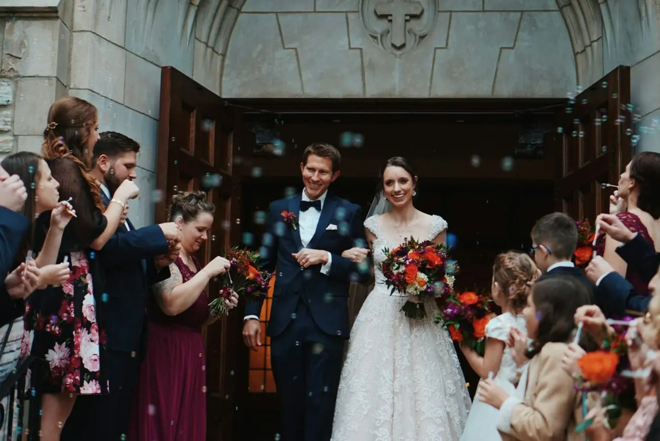 Sortie de cérémonie de mariage : les mariés heureux sont accueillis par des bulles de savon et leurs invités. La mariée porte une robe en dentelle et le marié un costume bleu marine. Scène festive avant le vin d'honneur.