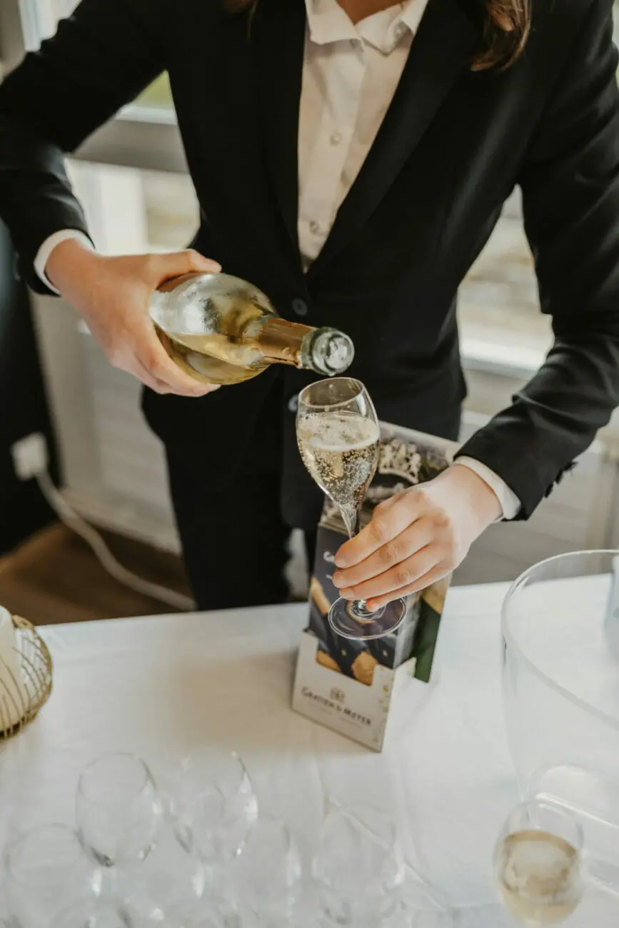 Une serveuse en veste de costume noire verse avec précision du vin pétillant d'une bouteille dans une flûte sur une table avec nappe blanche lors d'une réception de mariage.