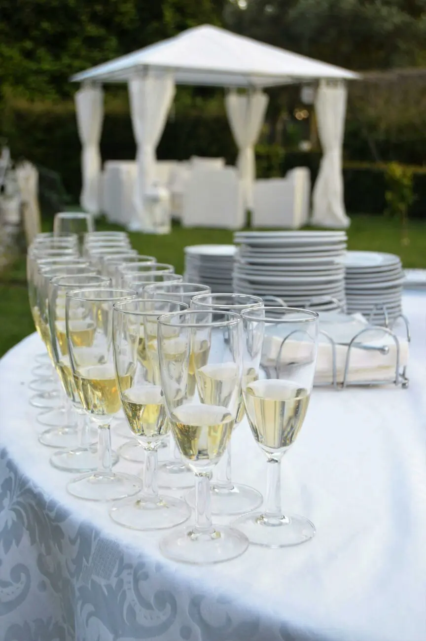 Gros plan de flûtes de champagne remplies et alignées sur une table de buffet avec nappe damassée, prête pour un vin d'honneur de mariage en extérieur. En arrière-plan, une tonnelle blanche élégante avec des voilages sur une pelouse. L'ambiance est festive et raffinée.