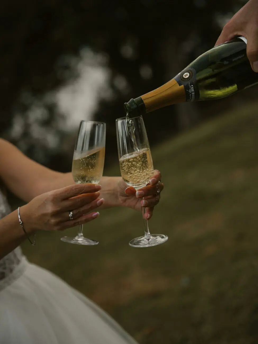Service de bulles dans deux flûtes lors d'une réception de mariage en extérieur, focus sur la robe dorée et les bulles.