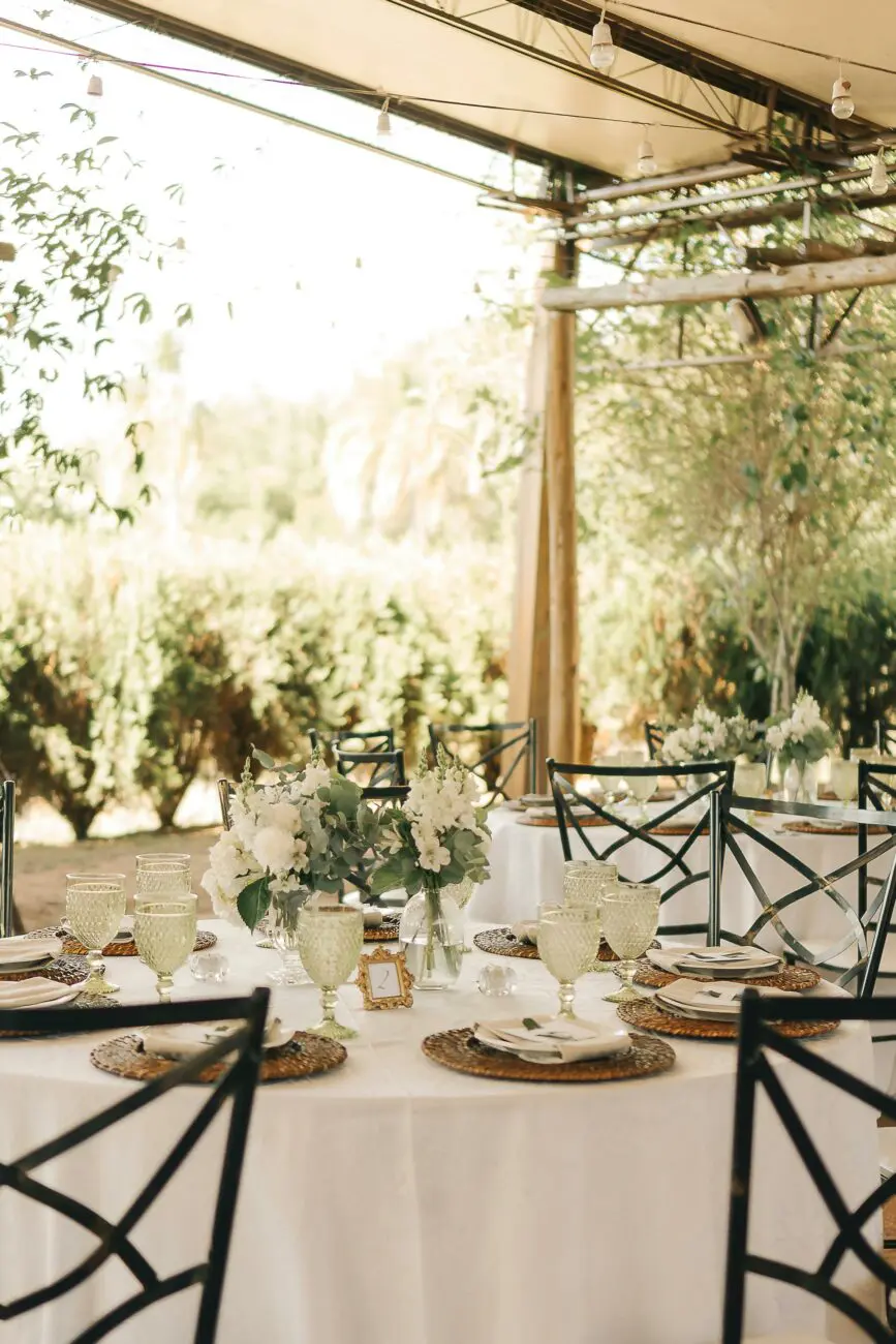Vue d'une table de mariage rectangulaire sous une structure couverte avec guirlandes lumineuses de type guinguette. Décoration de table épurée avec chaises en fer forgé.