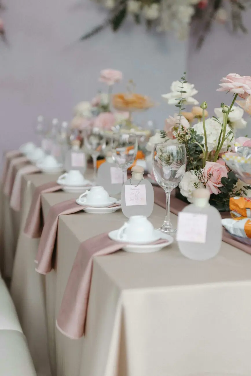 Table de mariage élégante avec nappage beige, fleurs roses et petits flacons de vin personnalisés pour les invités.