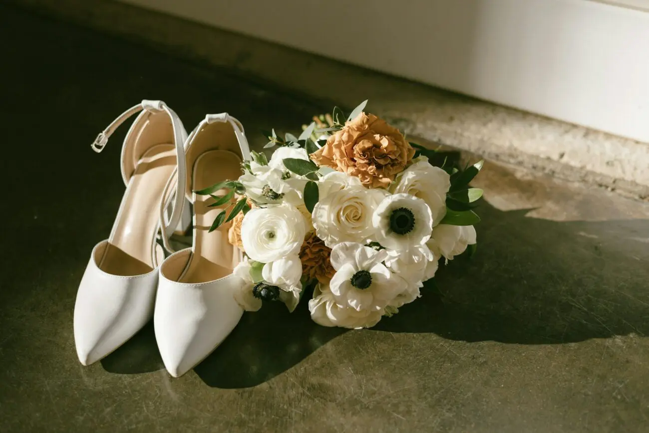 Accessoires de la mariée : chaussures de mariage blanches à talons et bouquet de fleurs avec anémones blanches et roses ocre sur un sol en béton ciré baigné de lumière.