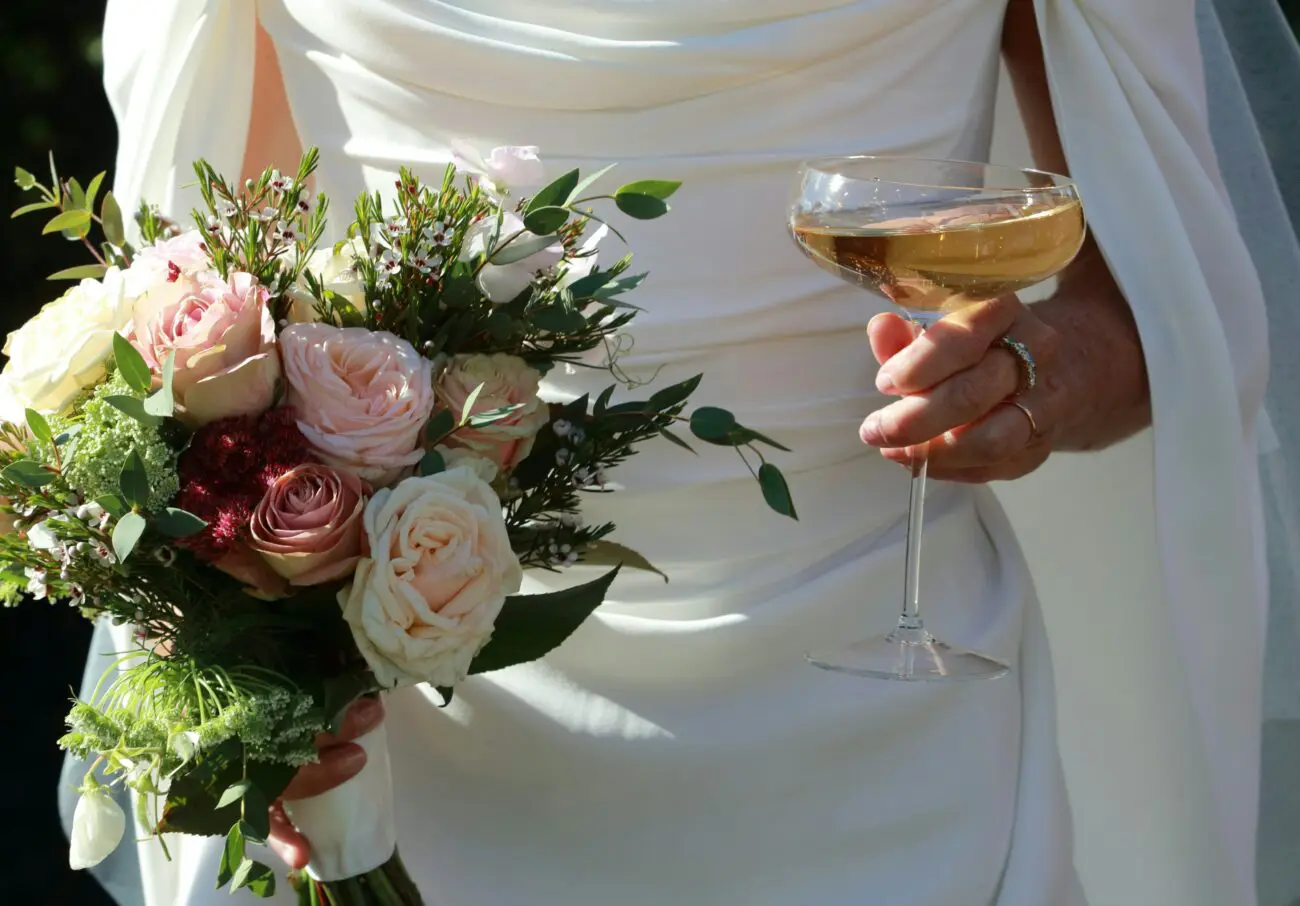 Gros plan sur une mariée en robe blanche tenant un bouquet de roses pastel dans une main et une coupe de champagne de style vintage dans l'autre.