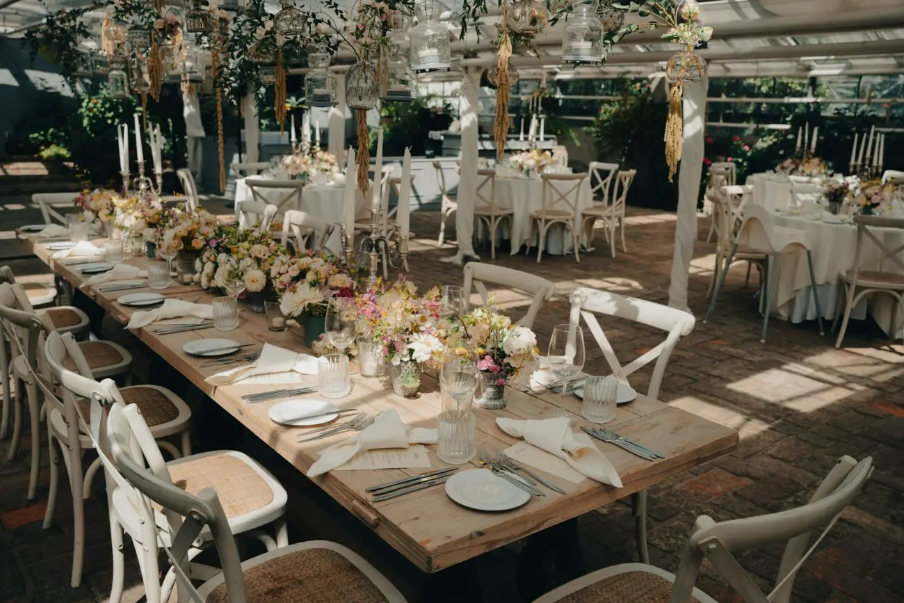 Grande table de banquet en bois sous une verrière lumineuse pour un mariage. Décoration florale abondante, chaises croisées blanches et verres à vin pour une réception élégante en intérieur.