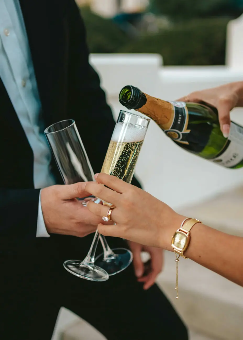 Gros plan sur les mains de jeunes mariés ou d'un couple de fiancés, avec bague de fiançailles et montre dorée, tenant des flûtes. Une main verse du champagne brut.