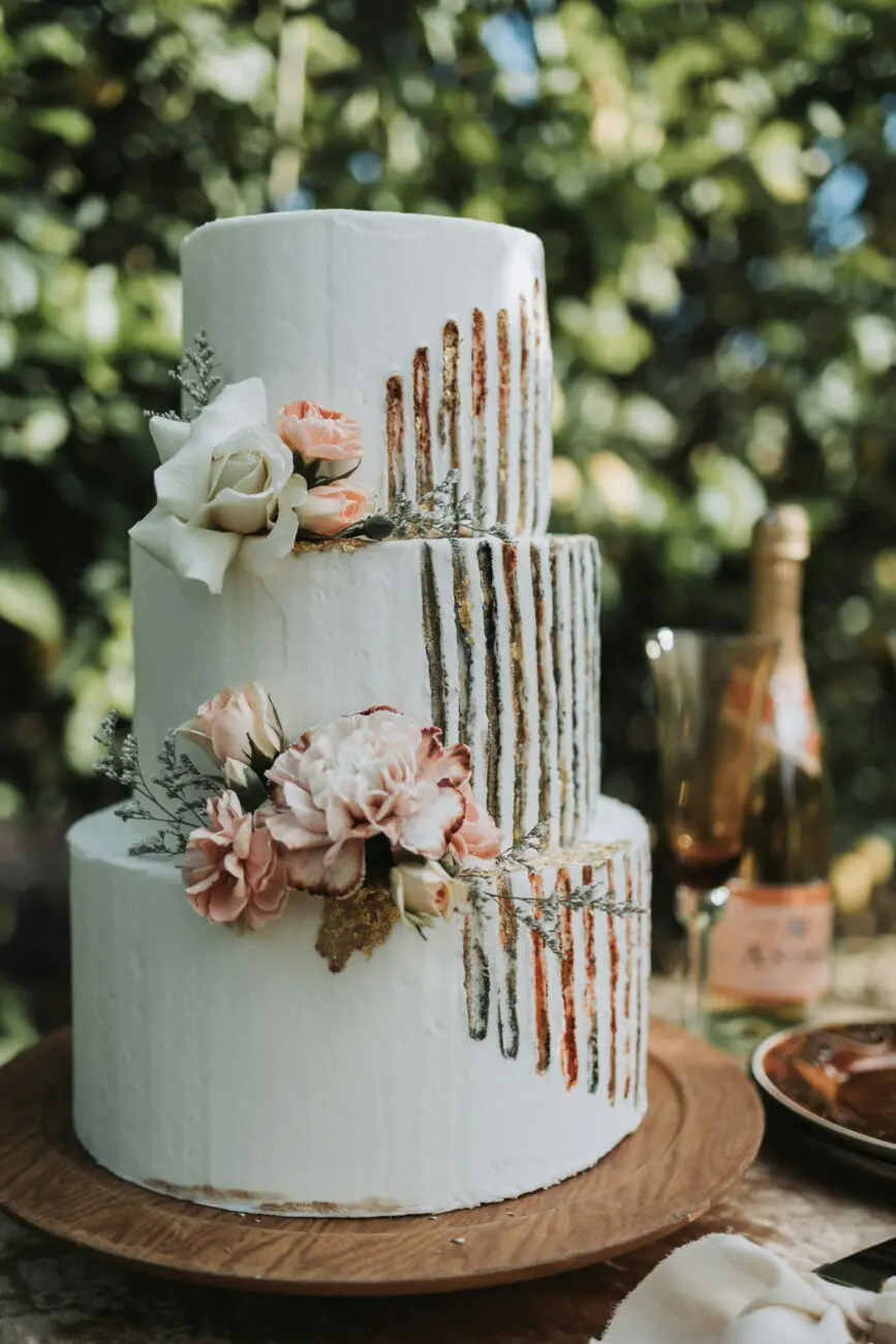 Gâteau de mariage blanc à trois étages avec fleurs pèche et dorures, accompagné d'une bouteille de champagne rosé en arrière-plan.