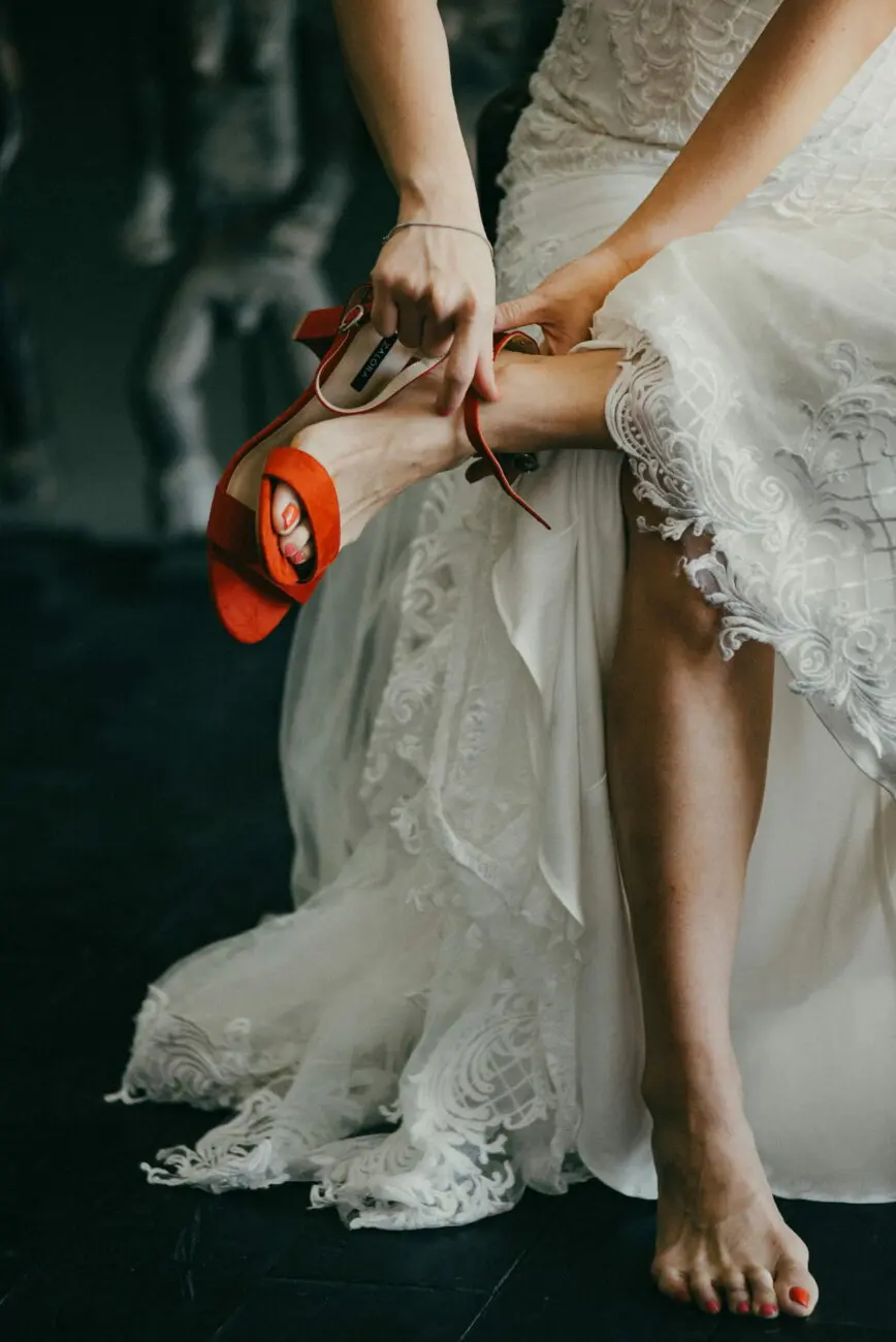La mariée ajuste une de ses chaussures à talons rouges, contrastant avec sa robe de mariée en dentelle blanche.