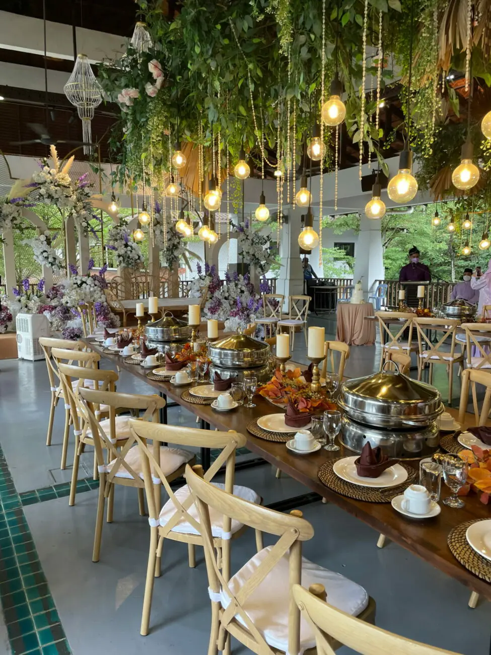 Grande salle de réception décorée de plantes tombantes et d'ampoules suspendues pour un buffet de brunch de lendemain de mariage, suggérant une sélection de vins de vins-mariages.net.