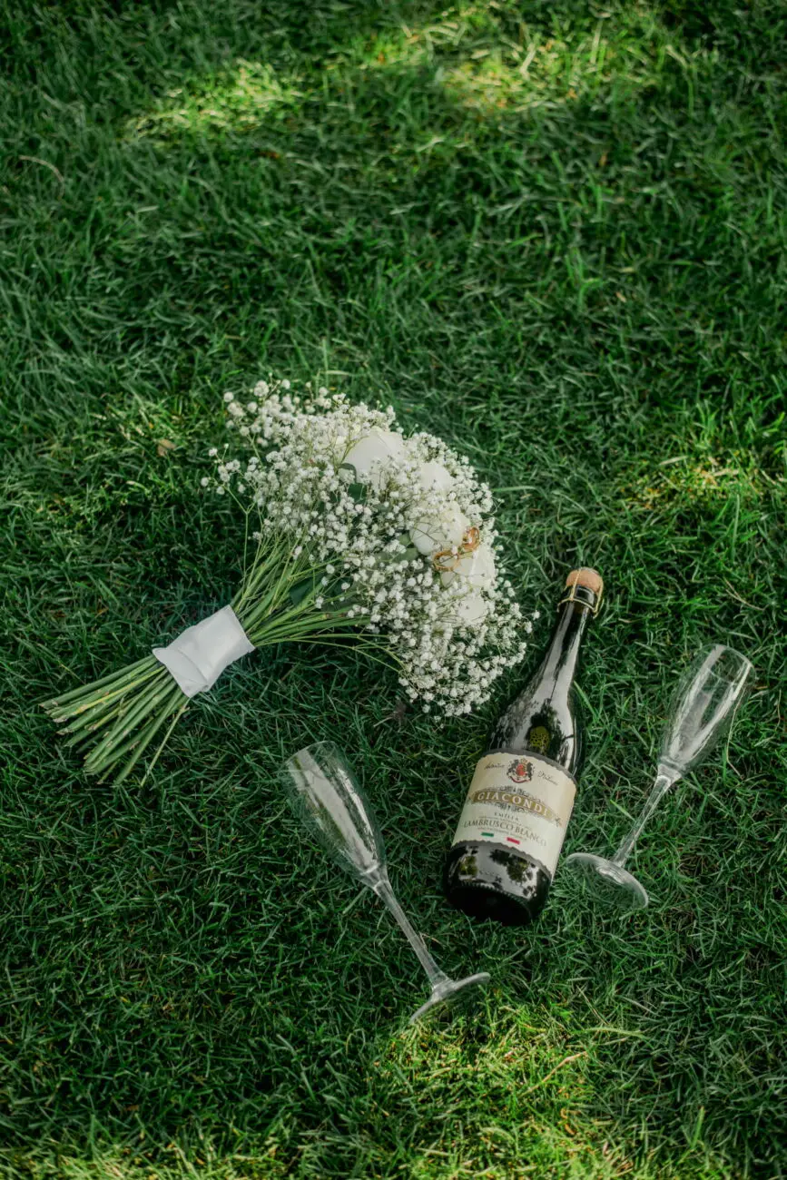 Un bouquet de mariée en gypsophile blanche posé sur l'herbe à côté d'une bouteille de Lambrusco Bianco et de deux flûtes à champagne vides.