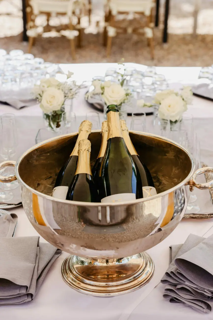 Grande vasque à champagne en métal argenté contenant plusieurs bouteilles au frais sur une table de mariage avec nappage blanc et roses blanches.