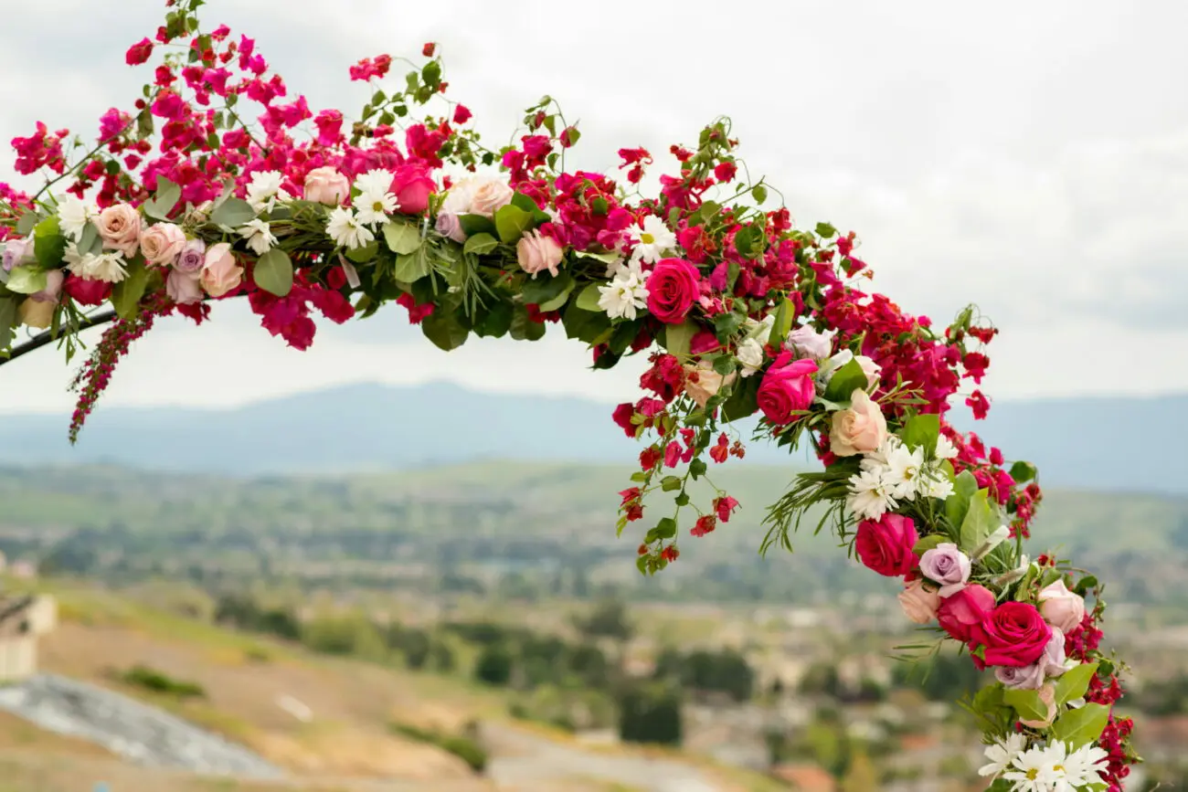 Arche de mariage décorée de fleurs roses intenses, blanches et mauves sur fond de paysage de collines verdoyantes.