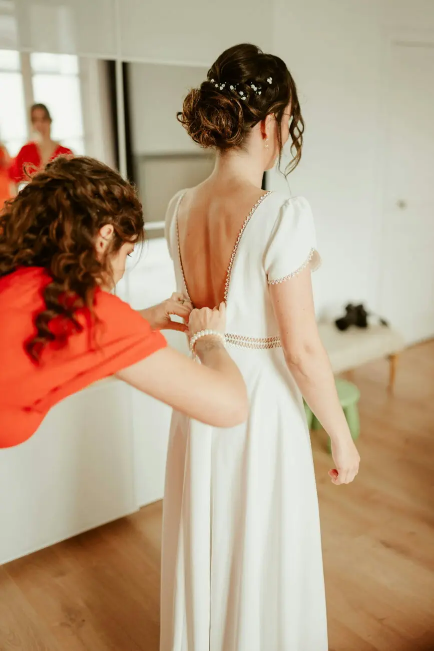 Séance d'habillage de la mariée dans sa robe blanche à dos nu, aidée par une amie, lors des préparatifs du mariage.
