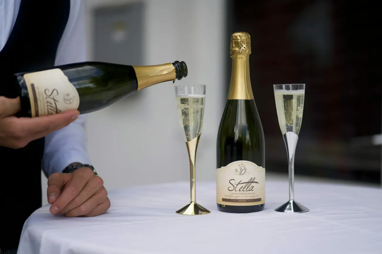 Un serveur verse du vin pétillant d'une bouteille dans une flûte dorée sur une table nappée de blanc, à côté d'une bouteille fermée et d'une flûte argentée.