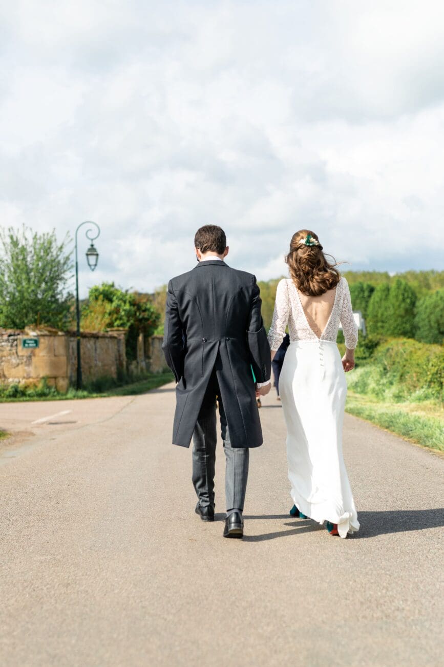 Couple de mariés élégants marchant sur une route de campagne en France après leur cérémonie, main dans la main. L'image illustre un moment de bonheur partagé, parfait pour une sélection de vins de mariage d'exception.