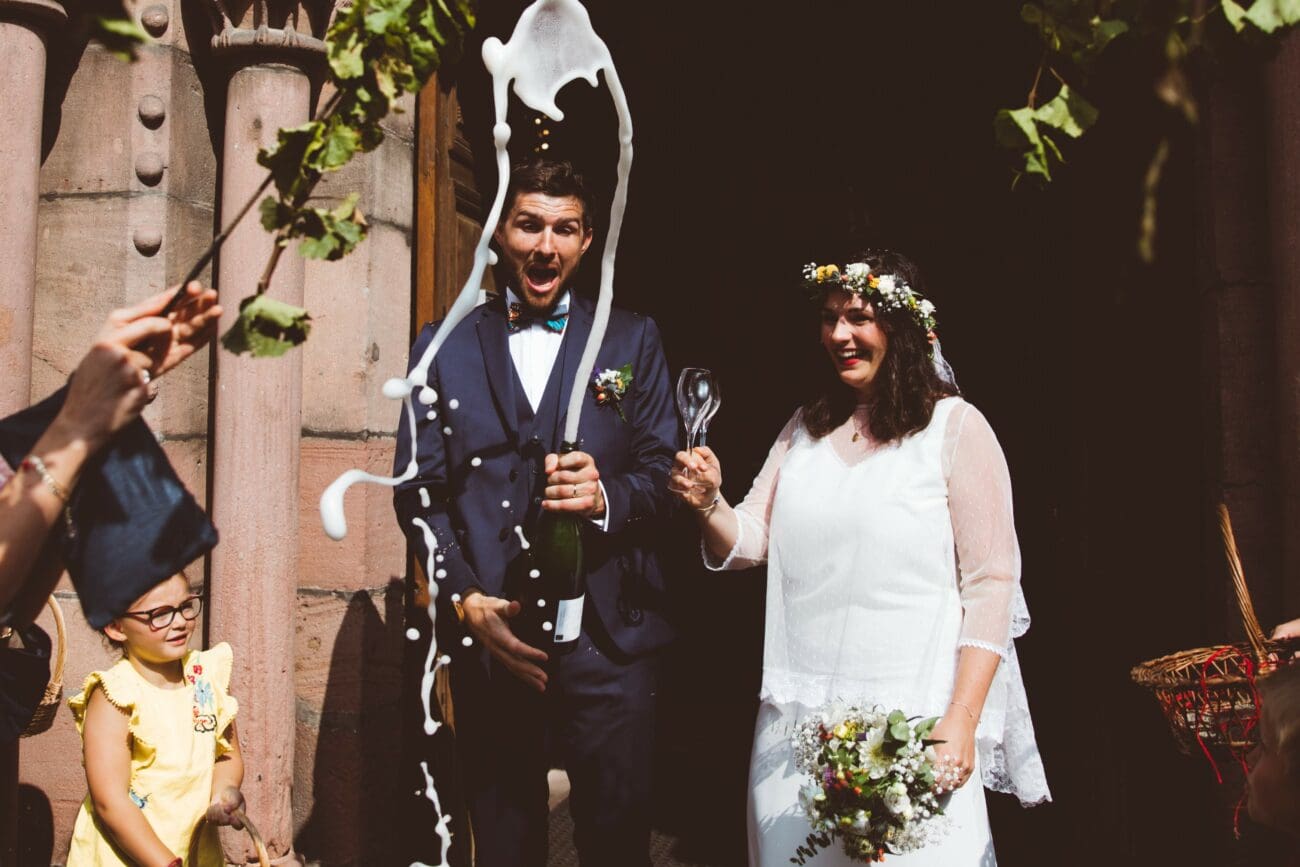 Marié ouvrant joyeusement une bouteille de Champagne avec un jet de mousse, aux côtés de la mariée tenant une flûte à la sortie de l'église.