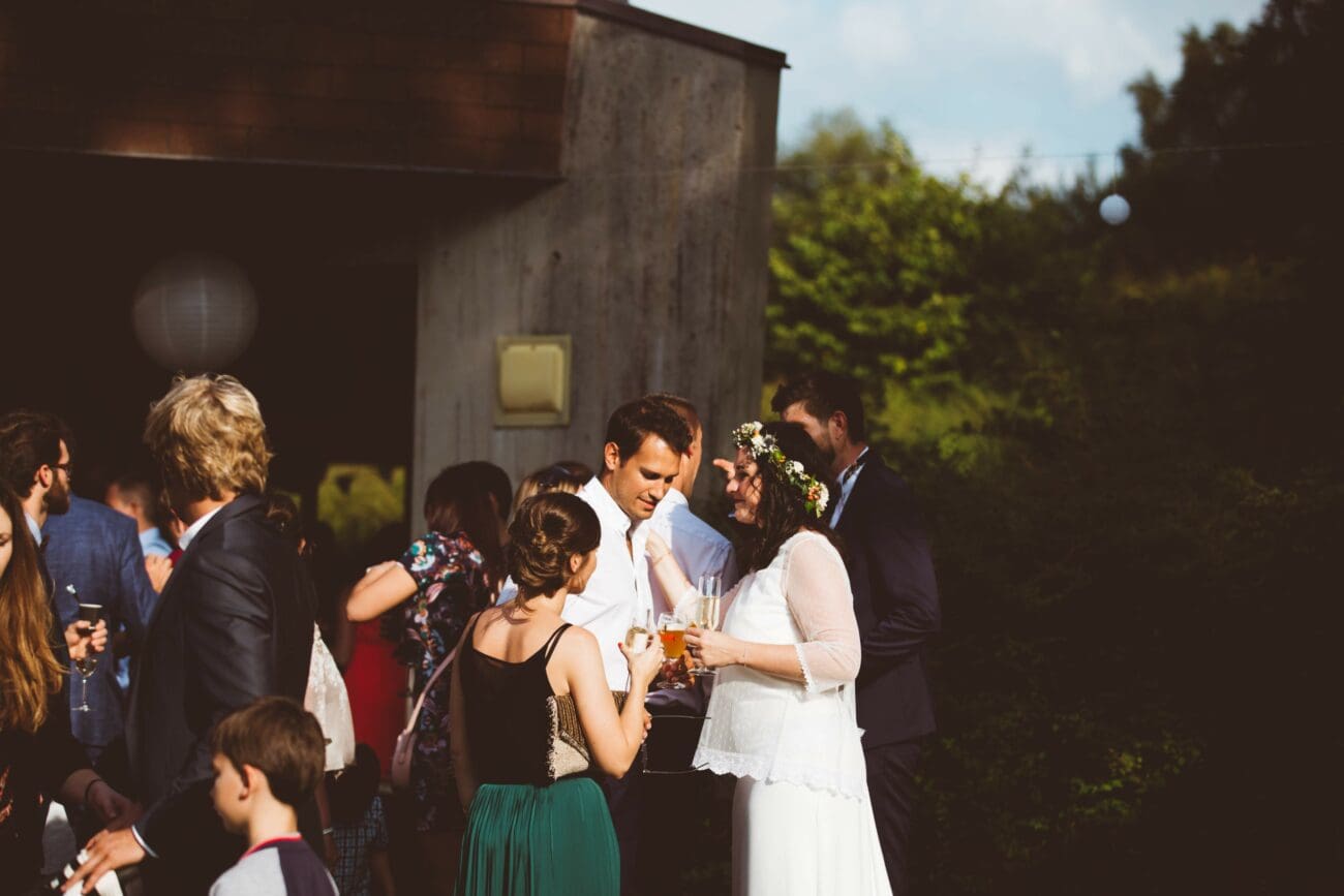 Un couple de jeunes mariés souriants, elle avec une couronne de fleurs, partageant un verre au milieu de leurs invités lors d'une réception.