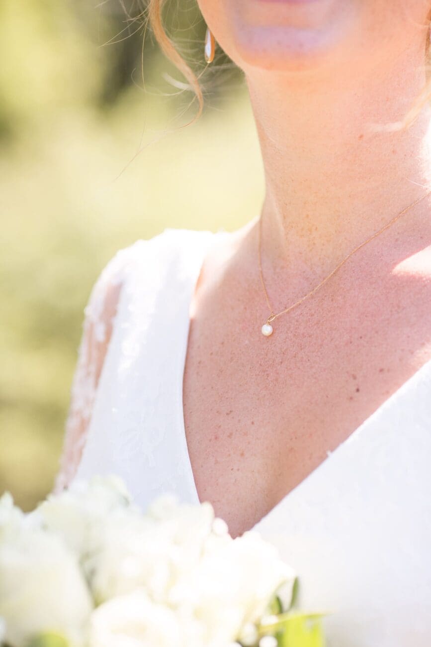 Gros plan délicat sur le décolleté d'une mariée, montrant un collier en fine chaîne d'or avec un pendentif en perle, une boucle d'oreille assortie et le haut d'un bouquet blanc flouté.