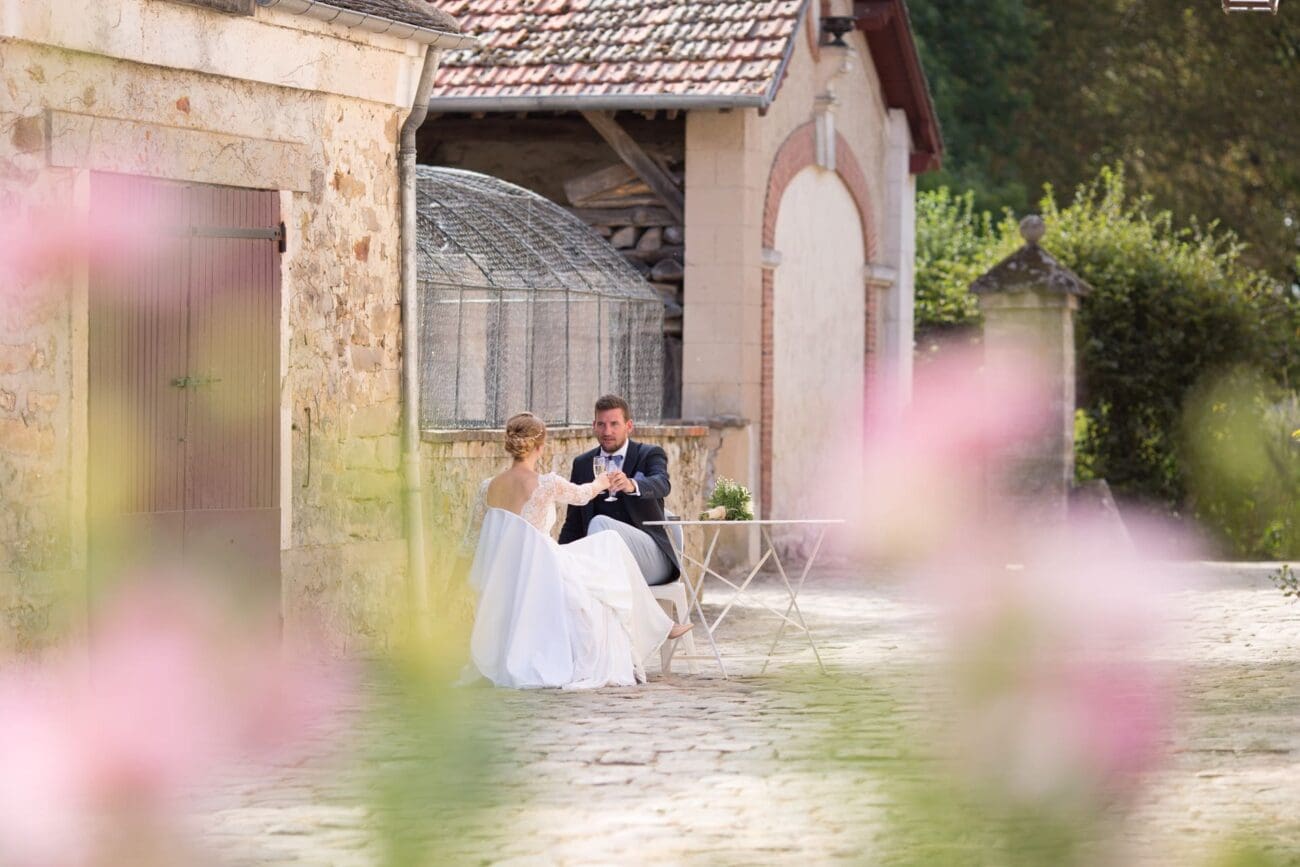 Vue de dos d'un couple de mariés assis à une petite table dans une allée pavée, entourée de bâtiments en pierre et de fleurs floues au premier plan, trinquant avec des flûtes.