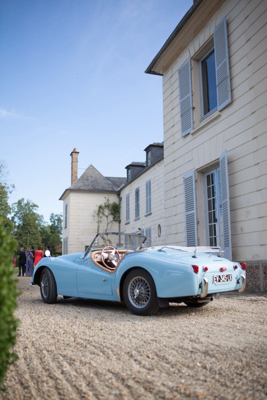 Une voiture décapotable bleu layette classique est garée sur une allée de gravier devant un élégant château à façade en pierre blanche. Des invités d'un mariage en tenue de cocktail sont visibles en arrière-plan, profitant de la journée ensoleillée.