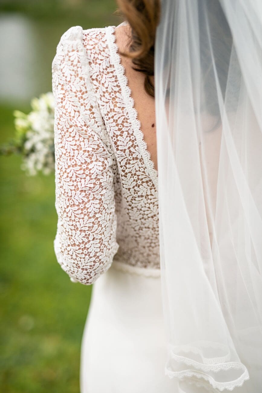 Détail de dos d'une mariée en robe de dentelle avec voile, tenant un bouquet en extérieur.