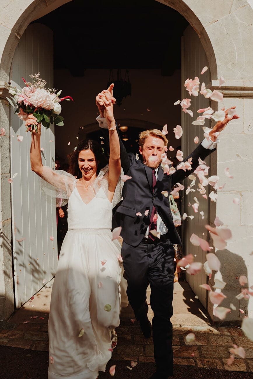 Mariés sortant de l'église sous un jet de pétales de roses roses, la mariée lève son bouquet et le marié exulte de joie.