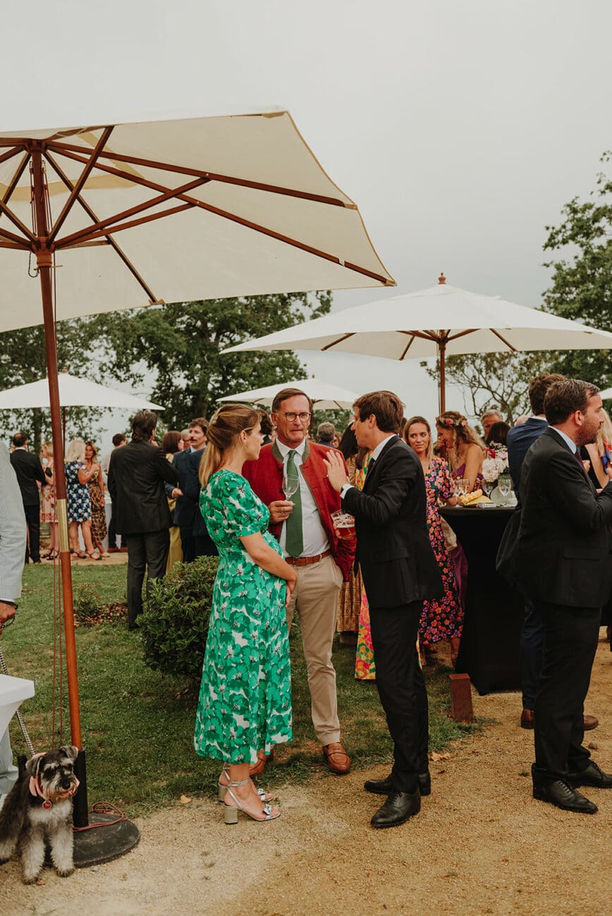 Invités d'un mariage discutant en extérieur sous des parasols blancs avec des verres de vin lors du cocktail.