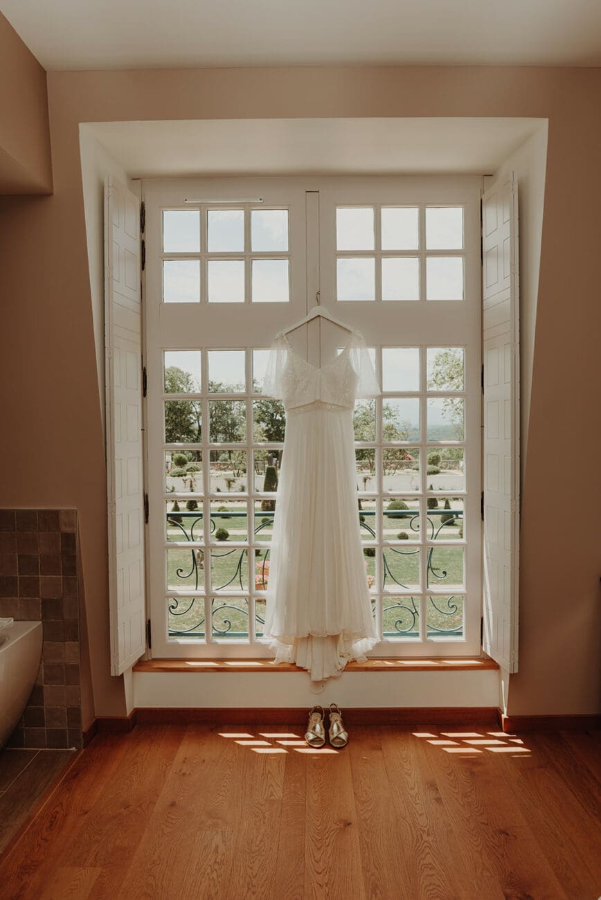 Une élégante robe de mariée en dentelle et mousseline est suspendue sur un cintre devant une grande porte-fenêtre à la française, donnant sur un jardin de château ensoleillé. Une paire de chaussures de mariée dorées est posée sur le parquet en bois en dessous.