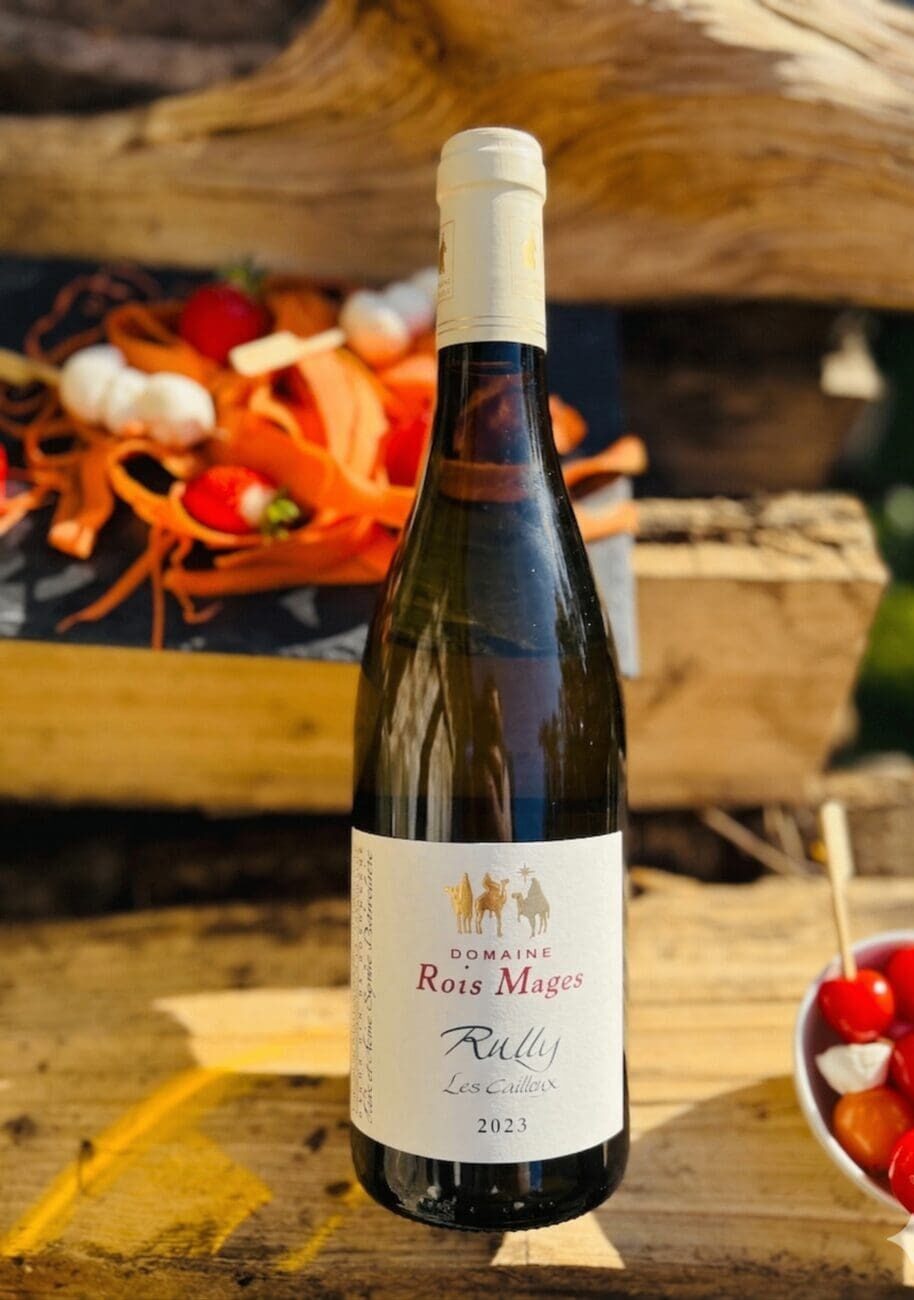 Bouteille de vin blanc Rully "Les Cailloux" 2023 du Domaine des Rois Mages posée sur une poutre en bois avec des amuses-bouches ensoleillés en arrière-plan.