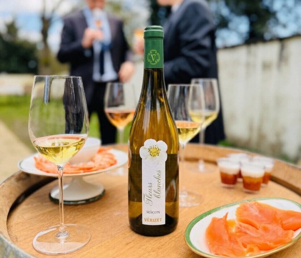 Bouteille de vin blanc Mâcon "Fleurs Blanches" 2024 de la Cave de Viré sur un tonneau lors d'un cocktail de mariage, entourée de verres de vin et d'assiettes de saumon et crevettes.