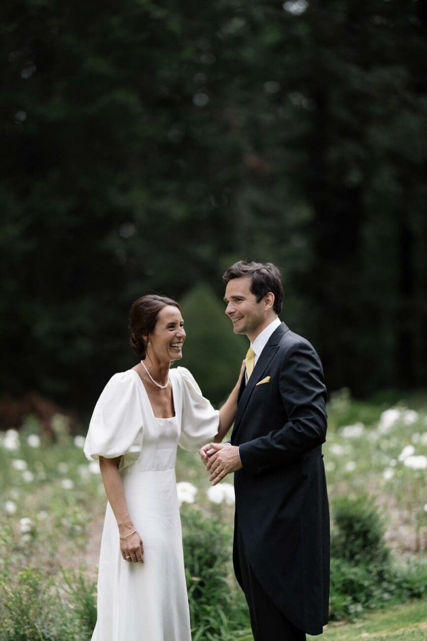 Portrait d'un couple de mariés rayonnants de bonheur dans un jardin. La mariée porte une robe blanche élégante à manches bouffantes et un collier de perles. Le marié est en jaquette sombre avec une cravate et une pochette jaunes. Ils sourient et se regardent avec complicité dans un cadre naturel verdoyant. Image illustrant la célébration parfaite d'un mariage.
