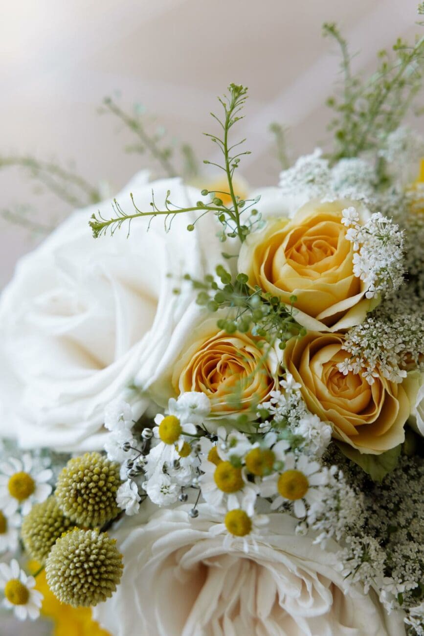 Gros plan artistique d'un bouquet de mariée aux teintes lumineuses, composé de grandes roses blanches, de roses jaune doré, de petites camomilles et de fleurs sauvages légères. L'image dégage une ambiance fraîche, champêtre et élégante, idéale pour inspirer une sélection de vins de mariage printaniers.