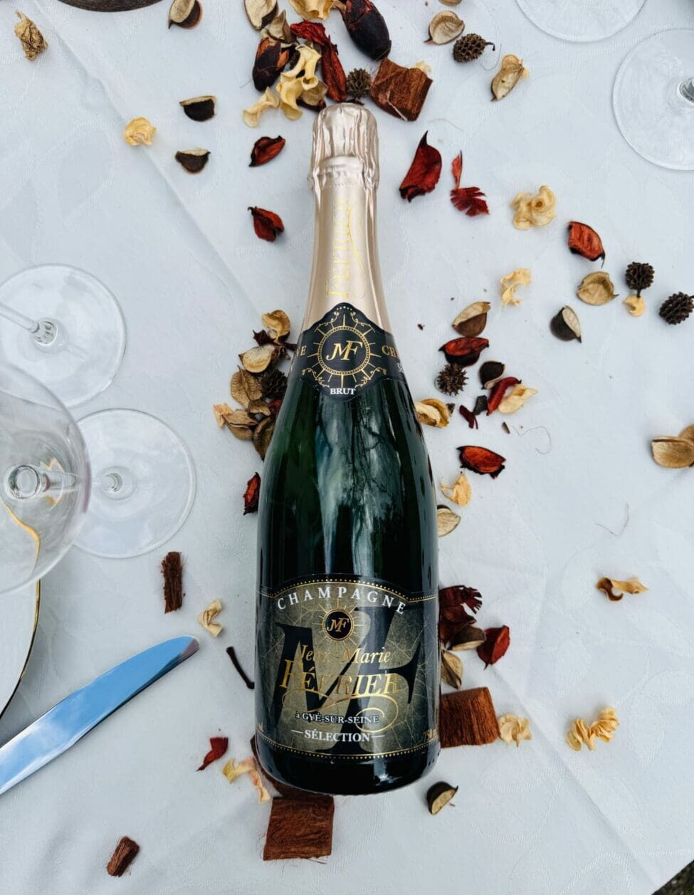 Vue d'en haut d'une bouteille de Champagne Jean-Marie Février Brut Sélection sur nappe blanche avec pétales séchés et couverts.