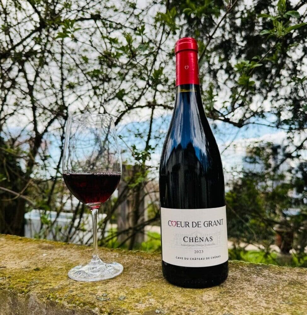 Bouteille deChénas Cœur de Granit 2023 Château de Chénas et un verre de vin rouge posés sur un muret en pierre en extérieur.