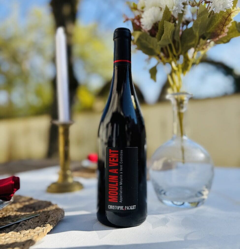 Bouteille de vin rouge Moulin-à-Vent 2023 de Christophe Pacalet posée sur un tonneau en bois avec une ardoise décorative et des fleurs jaunes.