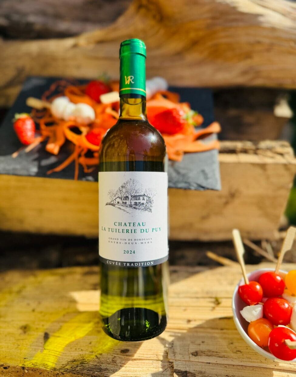 Bouteille de vin blanc Entre-deux-mers 2024 Château La Tuilerie Du Puy sur une caisse en bois avec des brochettes apéritives de tomates et mozzarella.