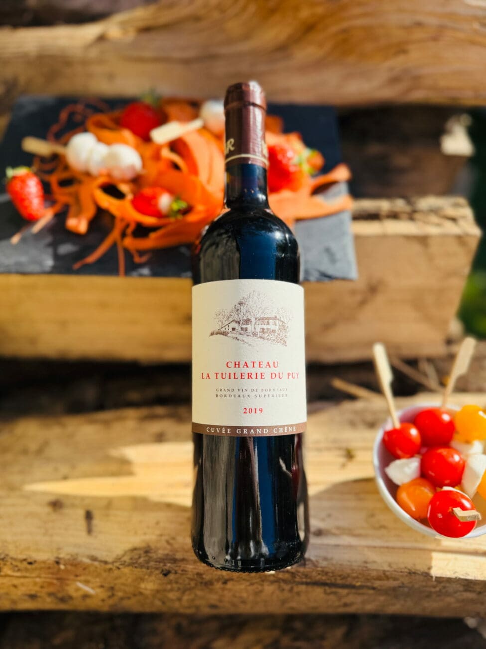 Bouteille de vin rouge Château La Tuilerie Du Puy "Cuvée Grand Chêne" 2019 posée sur des planches en bois lors d'un cocktail de mariage en extérieur, avec des amuses-bouches.