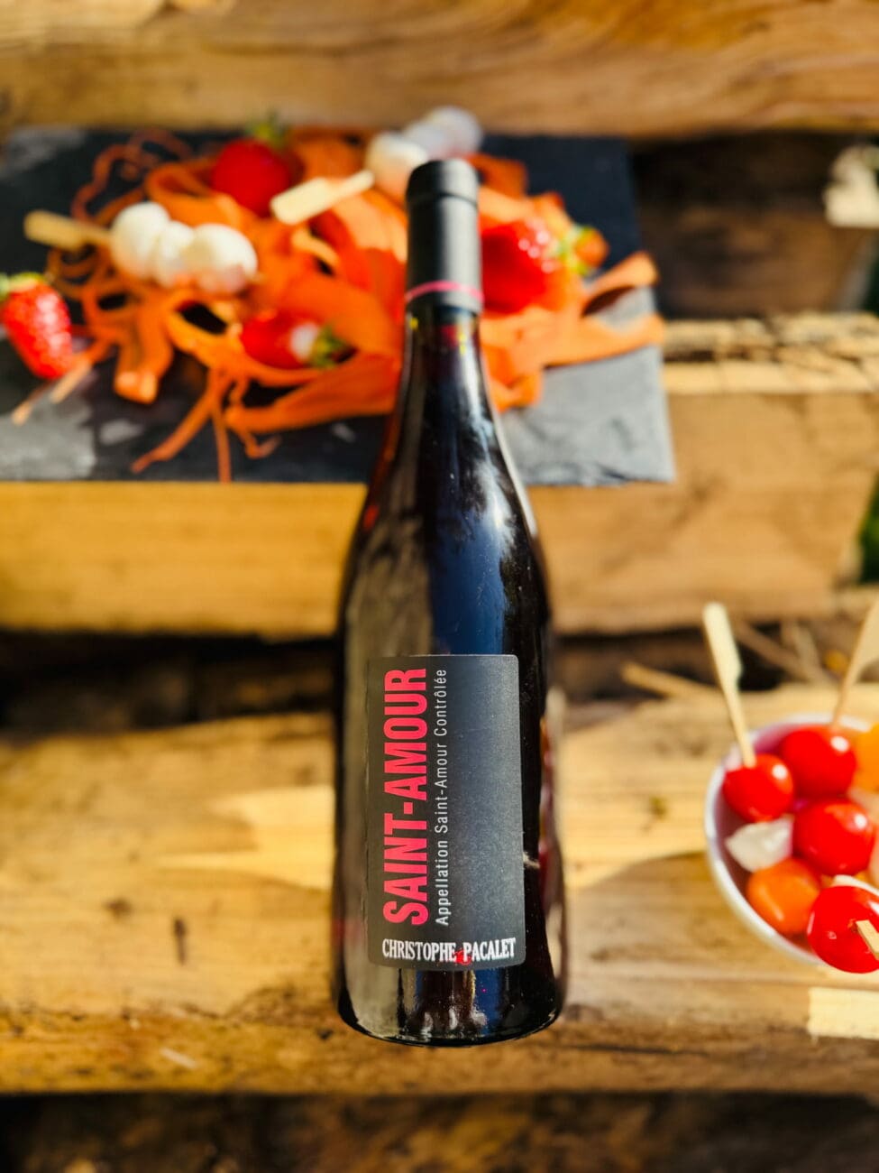 Bouteille de vin rouge Saint-Amour 2023 de Christophe Pacalet présentée sur une caisse en bois avec des accompagnements apéritifs colorés.