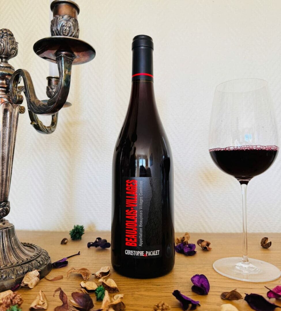 Bouteille de vin rouge Beaujolais-Villages 2023 de Christophe Pacalet avec un verre de vin servi à côté d'un bougeoir en métal argenté et des pétales séchés.